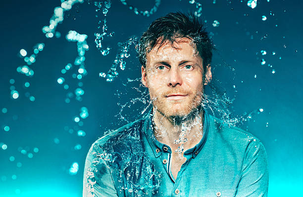Eyes wide open as man water in his head Portrait of a man who wears a shirt. He gets a bucket full of water in the head, but do not close his eyes. His shirt, hair and face gets wet. Lots of small drops. The photo is shot hispeed. The background is dark blue. slow motion stock pictures, royalty-free photos & images