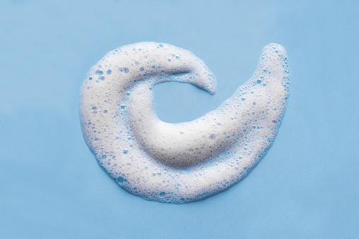 Face cleansing mousse swirl sample. White cleanser foam bubbles on blue background, copy space. Soap, shower gel, shampoo foam texture closeup.