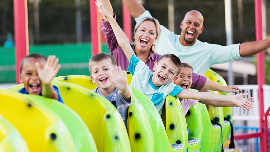 A multi-ethnic group of friends and family having run at an amusement park, on a colorful ride, a mini rollercoaster. The interracial couple are the parents of the two mixed race children, a 9 year old boy and 5 year old girl. The main focus is on their friend, the 9 year old boy in the middle. They are waving, laughing and shouting at the camera.