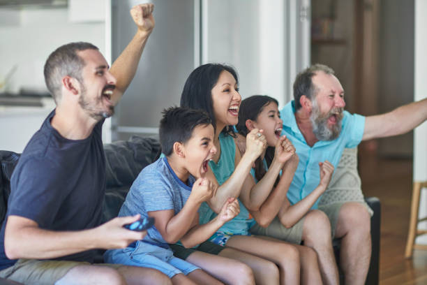 Family cheering while watching sport at home Multi-ethnic family cheering while watching sport at home. Enthusiastic fans are sitting on sofa while watching TV. They are in living room. asian kids watching tv stock pictures, royalty-free photos & images