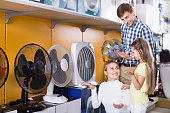 Smiling parents with daughter choosing cooling fan in home appliance store. Focus on woman and child