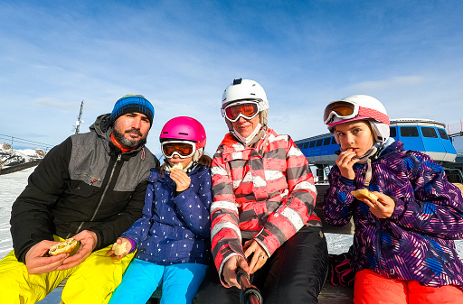 Family enjoying lunch on a break on vacations in skiing gear. Family with children on skiing vacation eating sandwiches in helmets and goggles. Winter sports mountain resort.