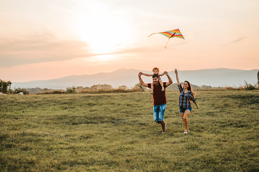 Young caucasian parents and their daughter running on a meadow with kite. Father carrying daughter on his shoulders.