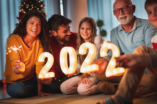 Happy family celebrating New Years Eve at home with kids, sitting by the Christmas tree, holding sparklers and illuminative numbers 2022 representing the upcoming New Year