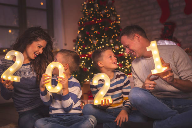 Family holding numbers 2021 Parents celebrating New Years Eve at home with kids, sitting by the Christmas tree, holding illuminative numbers 2021 representing the upcoming New Year happy new year 2021 stock pictures, royalty-free photos & images