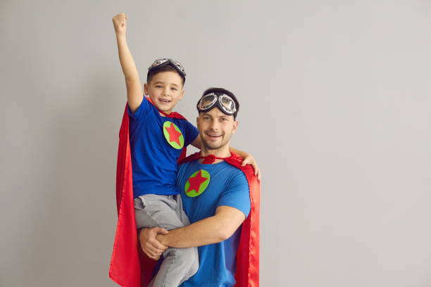 Family in superhero costumes. Father holds his son in his arms standing on a gray background. stock photo
