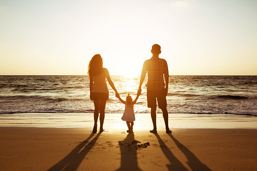 Family of father mother and daughter stands on background of sunset sea. Three silhouettes of happy people