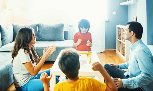 Family playing board game at home. Closeup of young family with two boys playing board game in their living room. They are seated on the floor around white table. One of the boys is on the move and the rest are cheering for him board game photos stock pictures, royalty-free photos & images