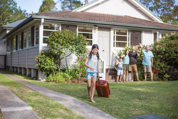 Family saying goodbye to girl leaving on trip Girl walking with luggage and family waving from background. Female exchange student in leaving house. They are wearing casuals in lawn. wave goodbye asian stock pictures, royalty-free photos & images
