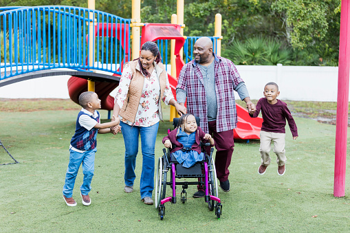 A happy African-American family with three young children walking together on a playground. The daughter, 5 years old, is in a wheelchair. She has caudal regression syndrome, a rare congenital disorder which affects the development of the lower spine. She is in the middle between the parents. Her brothers, twin 4 year old boys, are on the ends, holding their parents' hands.