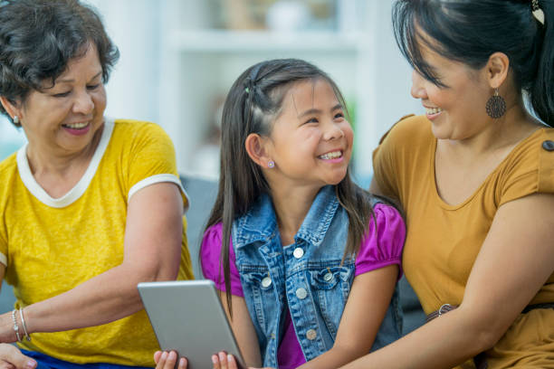 Family Watching A Video An Asian family is indoors in a living room. They are wearing casual clothing. A grandmother, mother and daughter are sitting on a couch and using a tablet. filipino family stock pictures, royalty-free photos & images