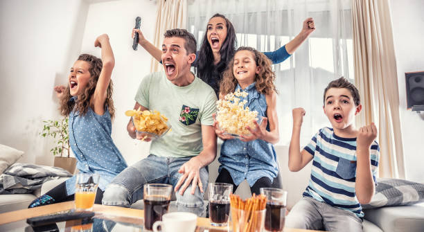 Family watching sports match on tv at home, cheering and shouting goal with hands up, spilling chips and popcorn from excitement. stock photo