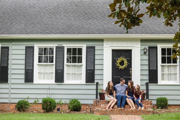 A family with a mother, father, and two daughters sitting outside on the brick steps of a front porch of a small blue cottage house stock photo