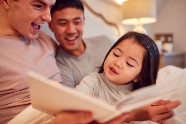 Family With Two Dads In Bed At Home Reading Story To Daughter stock photo