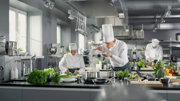 famous chef works in a big restaurant kitchen with his help. kitchen is full of food, vegetables and boiling dishes. he is trying taste. - chef imagens e fotografias de stock