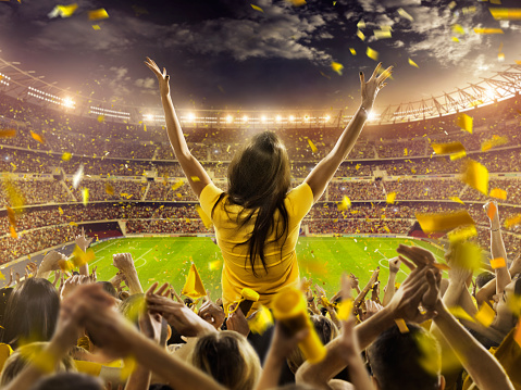 On the foreground a group of cheering fans watch a sport championship on stadium. One girl stands with his hands up to the sky. People are dressed in bright colors. A long-range shot of a stadium field, floodlights and seating. A green field, with painted white lines, is visible in the foreground. In the background are diffuse out-of-focus stadium seats. Large, bright floodlights are in the top-left and top-right corners of the image.