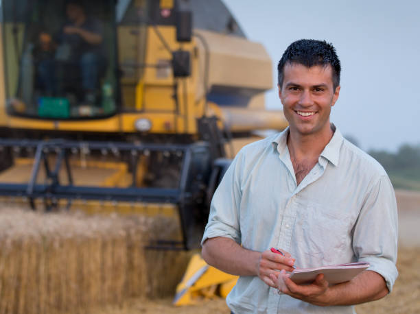 Farmer with notebook in front of combine harvester in field Happy young farmer engineer with notebook standing on wheat field while combine harvester working in background crop yield stock pictures, royalty-free photos & images