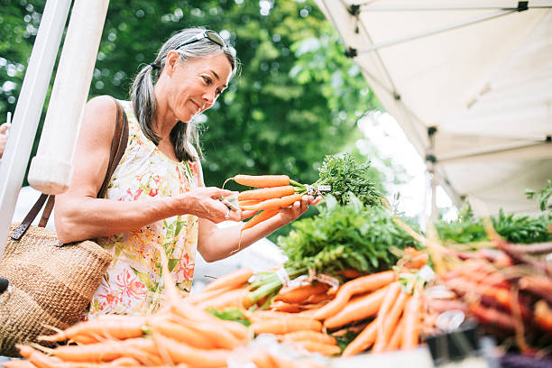 Farmers Market Shopping Mature Woman An older woman in her fifties shopping in a local farmers market with fresh, organic vegetables. She smiles as she chooses carrots. Horizontal image with copy space. farmer's market stock pictures, royalty-free photos & images
