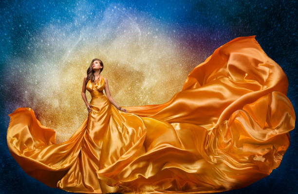 Fashion Model in Golden Dress over Miracle Night Sky Background. Luxury Woman in Long Silk Gown Dance and looking at Stars stock photo