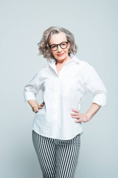 Fashion portrait of elderly businesswoman Elderly lady wearing white shirt, striped trousers and glasses standing with hands on hips against grey background, smiling at camera. Studio shot of female designer. hand on hip stock pictures, royalty-free photos & images