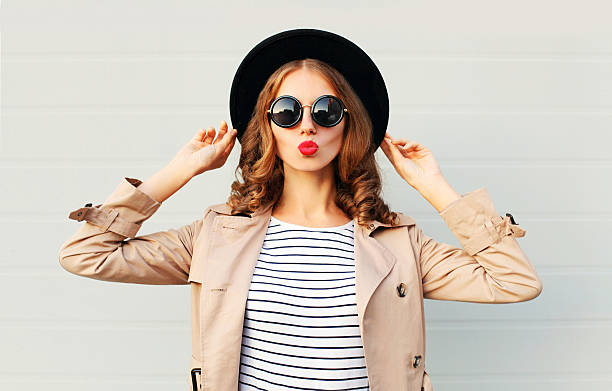 Fashion portrait pretty sweet woman blowing red lips, black hat Fashion portrait pretty sweet young woman blowing red lips wearing a black hat sunglasses coat over grey background fashionable stock pictures, royalty-free photos & images