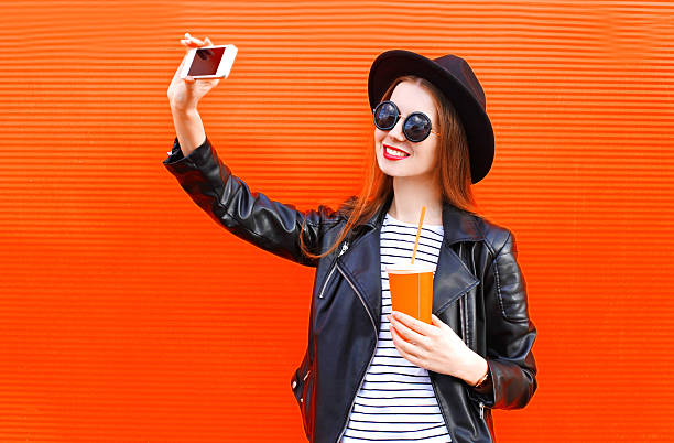 Fashion smiling young woman taking picture self portrait on smartphone Fashion pretty smiling young woman taking picture self portrait on smartphone in black rock style over city red background teenage girls photos stock pictures, royalty-free photos & images
