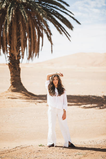 Fashion stylish portrait of young brunette woman in white shirt and pants. Palm, desert and sand dunes on background. Lifestyle concept. Fashion stylish portrait of young brunette woman in white shirt and pants. Palm, desert and sand dunes on background. Lifestyle concept. hot arabic girl stock pictures, royalty-free photos & images