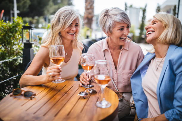 Fashionable senior friends drinking wine and having fun at home Happy mature women having fun and laughing together while drinking wine on restaurant patio wundervisuals stock pictures, royalty-free photos & images