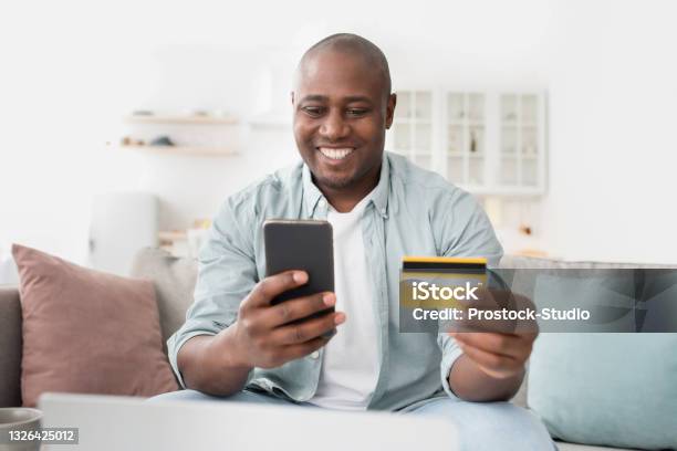 Fast online shopping. Cheerful african american man with credit card and smartphone sitting on couch at home