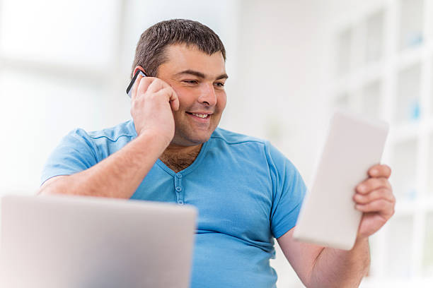 Fat man smiling and using wireless technology. Happy young overweight man talking on the cell phone and using digital tablet and laptop. fat man looks at the phone stock pictures, royalty-free photos & images
