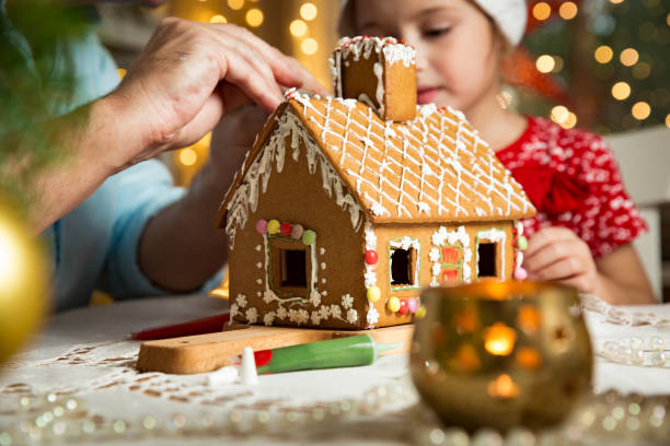 Father and adorable daughter in red hat building Christmas gingerbread house Father and adorable daughter in red hat building gingerbread house together. Beautiful decorated room with lights and Christmas tree, table with candles and lanterns. Happy family celebrating holiday. decorating stock pictures, royalty-free photos & images