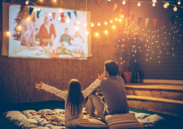 Father and daughter day. Father and daughter sitting at backyard and looking movie at home improved theatre. Backyard is decorated with string lighs. video sharing stock pictures, royalty-free photos & images