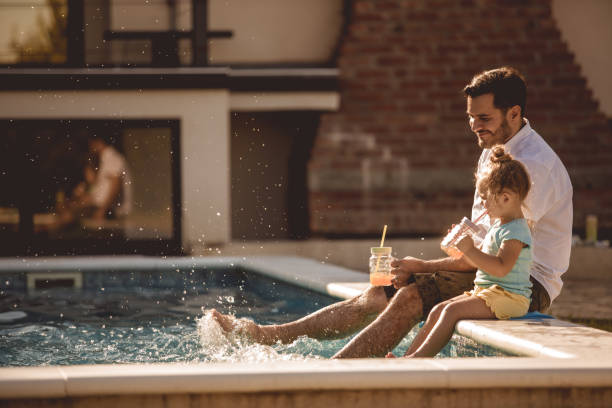 Father and daughter funny time by the pool Side view of lovely scene where daughter and dad plunge their feet in a pool while drinking fresh juices. airbnb stock pictures, royalty-free photos & images