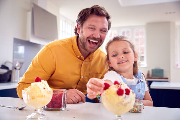 Father And Daughter In Kitchen Decorating Ice Cream Dessert With Cream And Raspberries stock photo