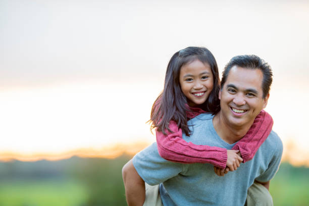 Father and daughter portrait outside in the summertime An asian elementary aged girl and holds onto her fathers back and smiles at the camera. Her father his smiling genuinely as he holds his daughter on his back. filipino family stock pictures, royalty-free photos & images