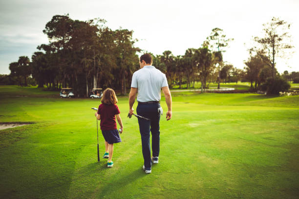 Father and daughter walk on a scenic golf course together, playing a round of golf with family stock photo
