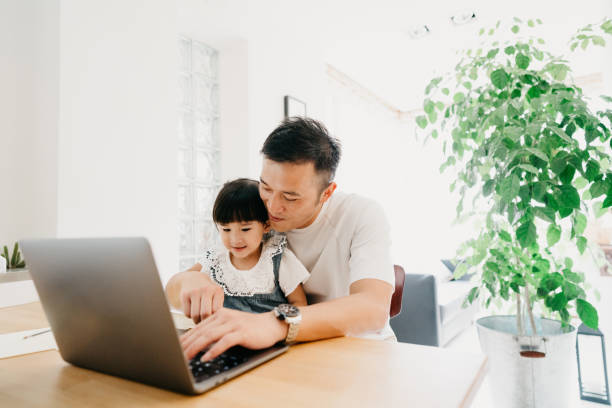 Father and daughter working together at home - The girl is following an online school lesson Father and daughter working together at home - The girl is following an online school lesson. They are at home during the Coronavirus Covid-19 pandemic. east asian culture stock pictures, royalty-free photos & images