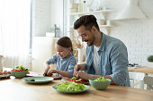 Father and small daughter cooking together, cutting vegetables fresh cucumber lettuce tomato and paprika for vegetarian salad. Healthy eating and lifestyle, teach develop kid, happy fatherhood concept