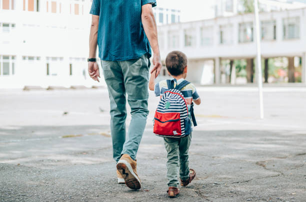 小さな男の子を学校に手をつないで導く父親の背面図。校庭を歩いているバックパックを持つ父と息子。