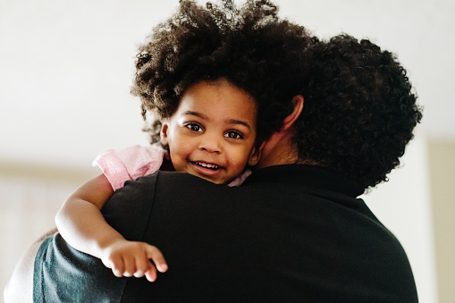 A mixed race father holds his black daughter while she smiles looking towards the camera.