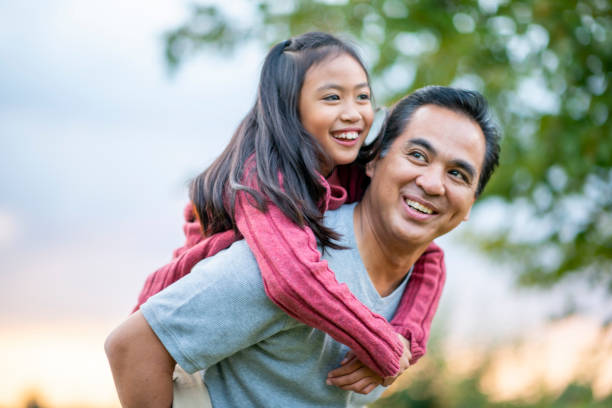 Father Giving His Daughter a Piggyback Ride stock photo A Filipino father gives his elementary school aged daughter a piggy back ride, while they enjoy a walk outside.  They are smiling and enjoying each others company and his daughter is looking down at him. filipino family stock pictures, royalty-free photos & images