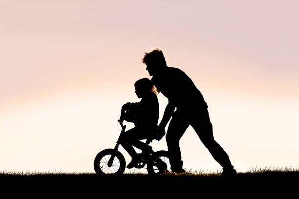 Father Helping his Young Child Learn to Ride Bike with Training Wheels A Silhouette of a good father is helping his young girl child learn to ride her bicycle with training wheels on a summer day. fathers day stock pictures, royalty-free photos & images