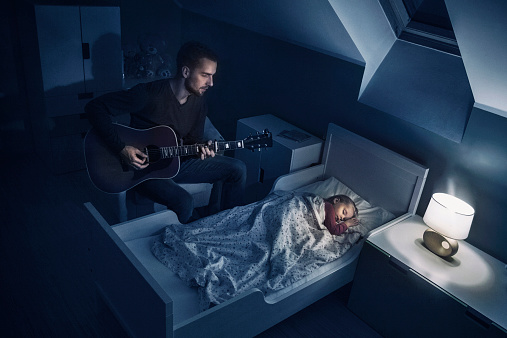 Photo of a father playing the guitar, singing lullaby for his sleeping daughter.