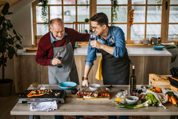Father teaching his son cooking tricks Senior father and young adult son preparing family lunch together in domestic home kitchen. Drink wine and have fun fathers day stock pictures, royalty-free photos & images