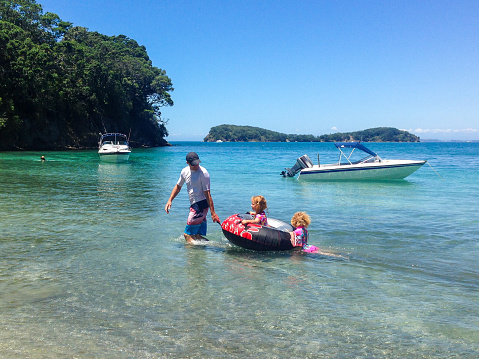 A father and two children at the beach, girls being towed on a rubber ring, Dairy Bay, Mahurangi Regional Park. Saddle Island and two boats in the background, Auckland, New Zealand