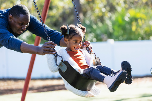 A father and daughter having fun together on the playground. The little girl, 3 years old, is sitting on a swing and dad is standing behind her, pushing, smiling and looking at her. The child is mixed race Hispanic, African-American and Native American.