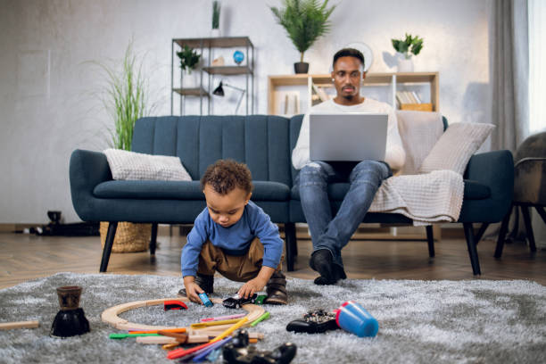 padre che lavora sul laptop mentre il figlio gioca sul pavimento - lavorare da casa foto e immagini stock