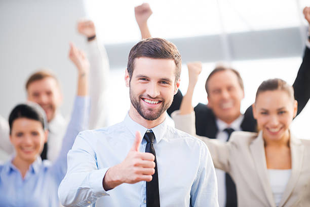 Feeling confident in his team. Happy businessman showing his thumb up and smiling while his colleagues standing in the background business thumbs up stock pictures, royalty-free photos & images