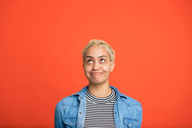 Young woman pulling a face smiling and looking up in front of an orange studio background.