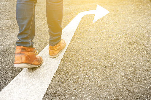 Feet and arrow on asphalt road background in starting line beginning idea. Selfie above view of hipster in boots or brown shoe standing on pathway. Top view. Moving forward, new start and success.